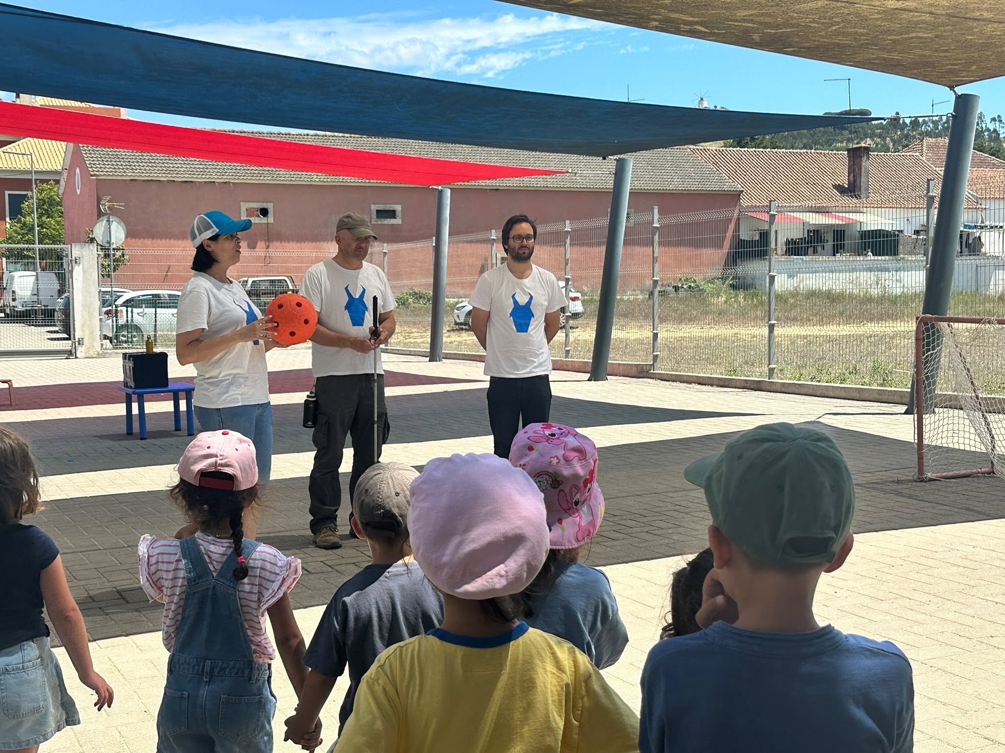 Crianças pequenas de costas, em fila, numa atividade ao ar livre. À frente delas, mais ao fundo, estão três adultos alinhados, voltados para as crianças. Todos os três usam camisola branca com o mesmo logótipo azul ao centro, que parece a silhueta estilizada de uma cabra. A pessoa à esquerda é uma mulher com cabelo escuro curto, boné azul e branco, calças de ganga claras e ténis. Nas mãos, segura uma bola laranja viva, de tamanho médio, com vários buracos redondos, conhecida por ser utilizada no desporto adaptado de goalball. Ao lado dela, ao centro, está um homem com barba curta grisalha, boné bege, calças escuras e botas, segurando uma bengala longa verticalmente em frente ao corpo. À direita está outro homem, com barba escura, óculos, calças escuras e ténis, com as mãos atrás das costas. Em primeiro plano, várias crianças pequenas estão de costas, viradas para os três adultos. Usam roupas de verão: t-shirts de cores variadas, jardineiras de ganga, calções e chapéus ou bonés rosa, verde, cinzento e estampados. Duas crianças de mãos dadas estão no centro; uma usa jardineira de ganga clara e boné rosa-claro, outra usa t-shirt amarela. A atividade decorre numa zona pavimentada coberta por toldos grandes de tecido esticado em azul-escuro, vermelho e bege, criando sombra. À direita há uma pequena baliza metálica de rede, parecendo de hóquei ou futebol de rua. No fundo da imagem vê-se uma vedação metálica transparente, alguns carros estacionados e edifícios baixos com telhados de telha, numa área de bairro residencial em dia ensolarado.